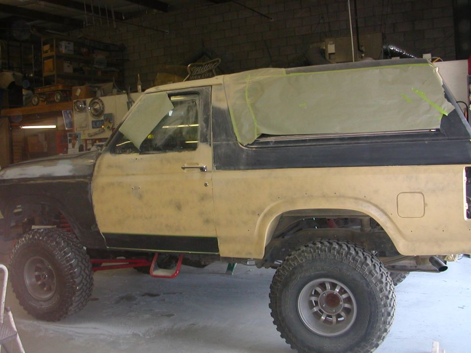 Full-size Ford Bronco in primer, hardtop masked with tape, lifted on mud tires in garage
