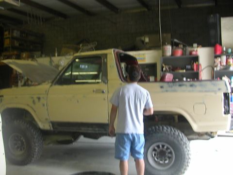 1984 Ford Bronco - A person stands beside a yellow Ford Bronco in a garage, with the hood open, indicating it is undergoing restoration.
