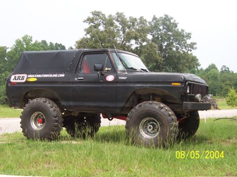 1978 Ford Bronco - Flat-black full-size Ford Bronco with a soft top, shown in profile on grass: heavily lifted on oversized mud tires and chrome wheels, red-p…