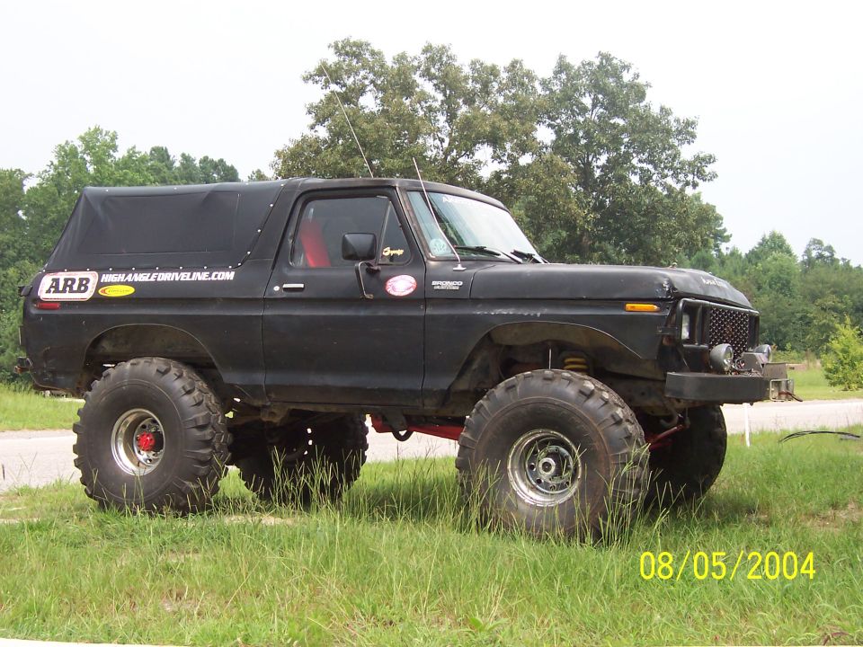 Lifted flat-black Ford Bronco with soft top and mud tires parked on grass, side view