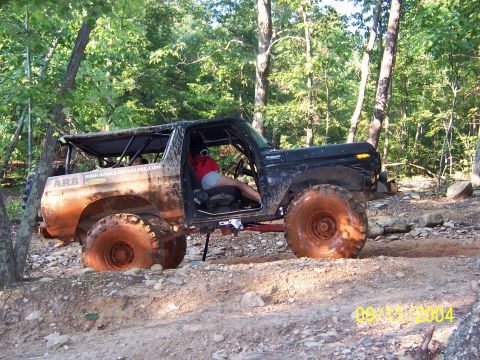 1978 Ford Bronco - Mud-caked black full-size Ford Bronco flexing over a rocky woodland trail, doorless and topless with an internal roll cage, ARB and HighAng…