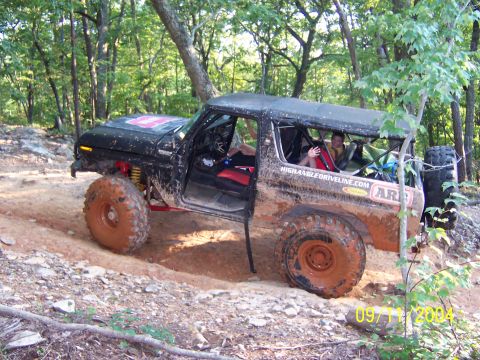 1978 Ford Bronco - Mud-caked black doorless full-size Bronco-style 4x4 flexing through a rutted red-clay trail in the woods, shot from the driver's side on 09…