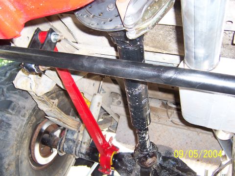 1978 Ford Bronco - Undercarriage view of a lifted 4x4 showing the rear driveshaft with a slip yoke and U-joint running to a solid axle, alongside a red-painte…