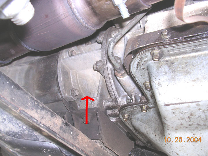 Underside view of vehicle transmission housing and exhaust pipe, with a red arrow pointing to a bolt. Date stamp reads 10.20.2004