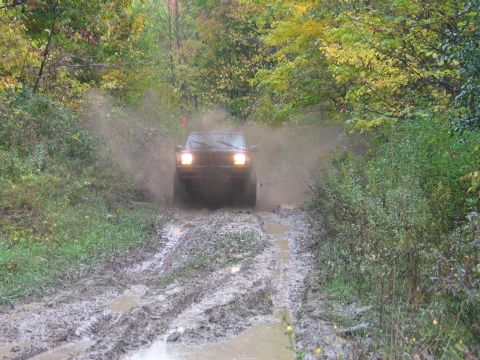 1995 Ford Bronco - A Jeep Cherokee XJ kicks up a large spray of mud as it navigates a waterlogged, rutted path through a forest.