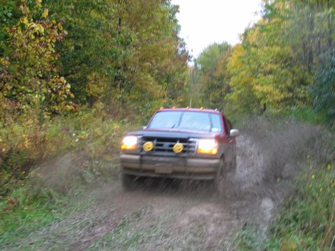 1995 Ford Bronco - A red pickup truck navigates a muddy trail, kicking up spray as it drives through the mire.