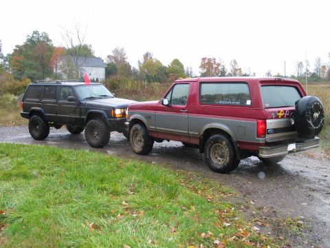 1995 Ford Bronco - Two SUVs, a 1995 Ford Bronco and a Jeep Cherokee, are parked on a dirt road.