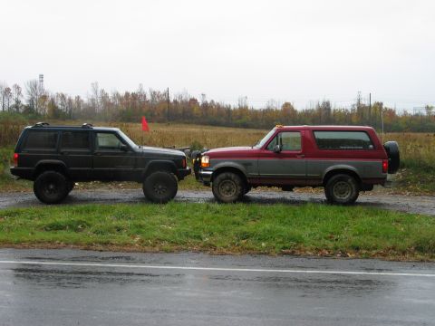 1995 Ford Bronco - Two off-road ready SUVs are parked side-by-side on a grassy shoulder next to a wet road.
