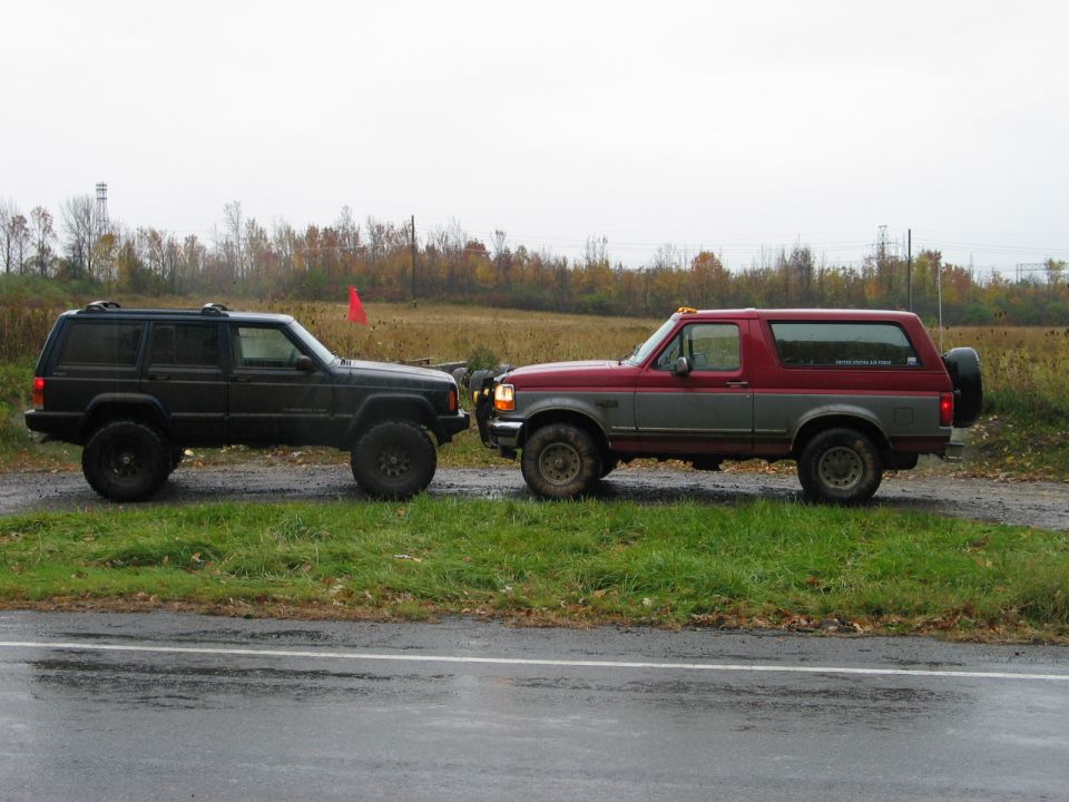 Red and gray lifted Ford Bronco parked beside a green lifted Jeep Cherokee on wet gravel