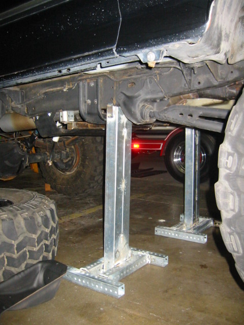 1992 Ford Bronco - A dark truck body sits raised on two tall galvanized slotted-steel stands placed under the frame rails, with the rear axle, leaf spring, mu…