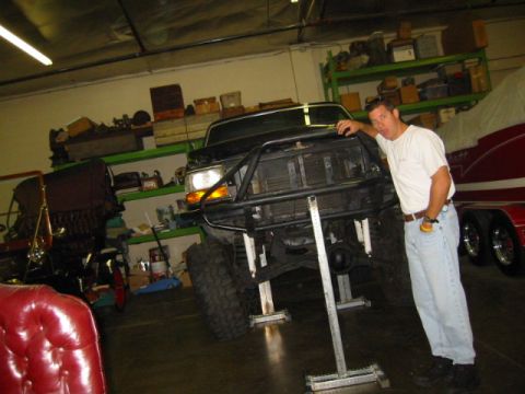 1992 Ford Bronco - A dark green Ford full-size truck/Bronco sits nose-up on tall adjustable jack stands in a garage, front fascia and grille removed to expose…
