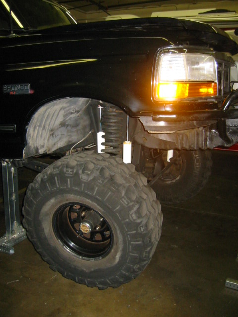 1992 Ford Bronco - Front three-quarter close-up of a lifted black Ford Bronco hanging on jack stands so the driver-side front suspension droops fully, showing…