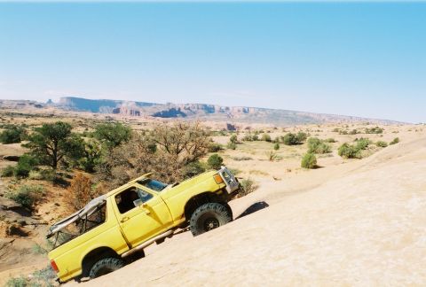 1996 Ford Bronco - A yellow SUV navigates a steep, sandy terrain under a clear blue sky.