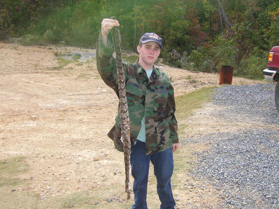 Guy in camo jacket and jeans holding up a long snake