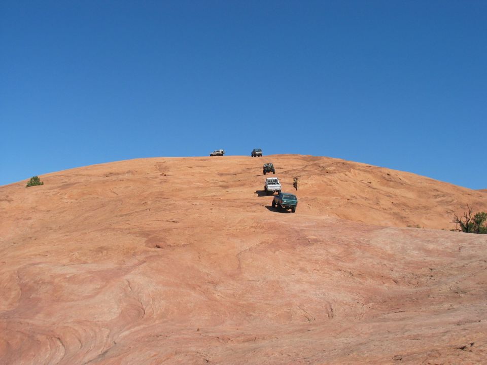 Several off-road vehicles ascend a large, red, rocky incline under a clear blue sky