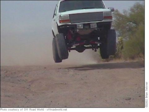 1993 Ford Bronco - A white SUV is captured mid-air during a jump over a dirt track, its lifted suspension and large tires.