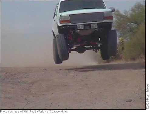 White SUV mid-air over a dirt track, front view, lifted suspension, large tires
