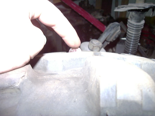 finger pointing at a coin on a metal engine component, with some hoses and a red bar in the background