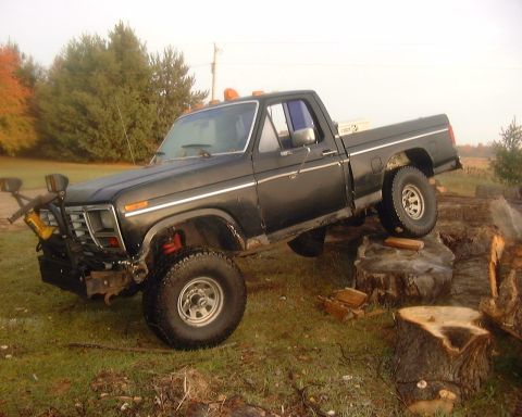 1985 Ford F-150 - A black Ford F-150 4x4 regular-cab pickup posed with its front passenger wheel climbed up onto a large tree stump, showing off its lifted s…