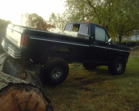 1985 Ford F-150 - This lifted black pickup truck is positioned on a large log, its side profile and a toolbox mounted in the bed.
