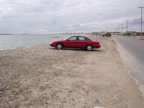 1998 Ford Taurus - A red 1998 Ford Taurus sits parked on a rocky shore beside a road with a distant SUV.
