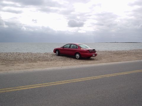 1998 Ford Taurus - A red 1998 Ford Taurus sits parked on a gravelly shore next to a calm ocean under a cloudy sky.