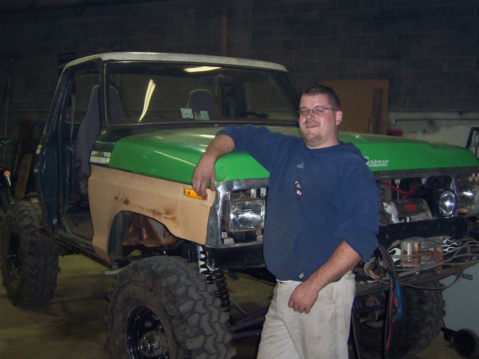 Man in blue sweater leaning on a green fender of a lifted truck under construction in a garage