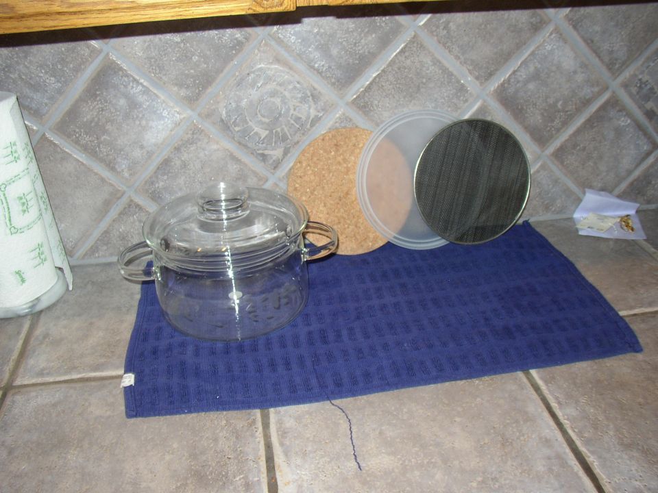 Glass pot with lid, cork coaster, and metal strainer on a blue towel on a tiled counter