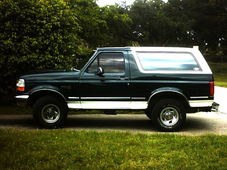 Side view of a dark green Ford Bronco SUV with white top parked on a driveway near trees