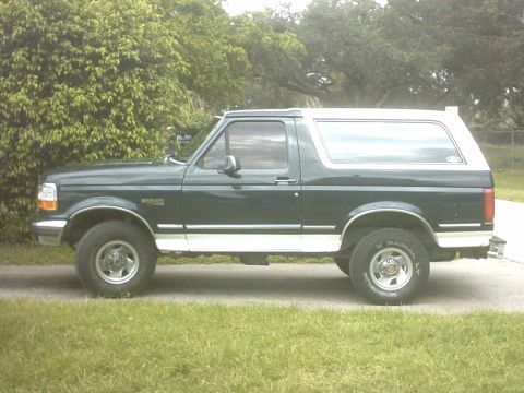 1992 Ford Bronco - Driver-side profile of a stock-height dark green Ford Bronco with a white removable hardtop, parked on a concrete driveway with trees and g…