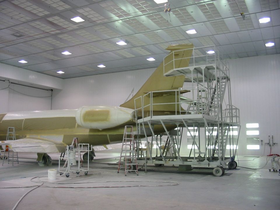 Private jet primed in yellow-tan paint on scaffolding inside a large white paint hangar