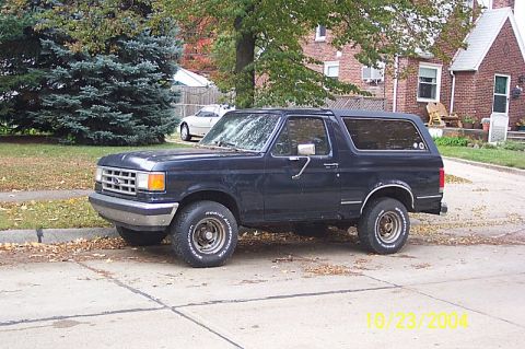 1978 Ford Bronco - A dark blue 1987 Ford Bronco is parked on a street in front of a house and trees.