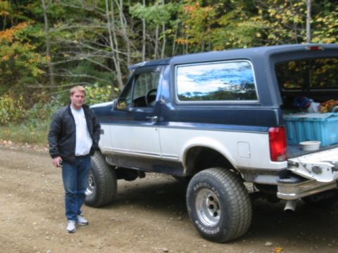 1996 Ford Bronco - A man stands beside a two-tone Ford Bronco parked on a dirt road with trees in the background.