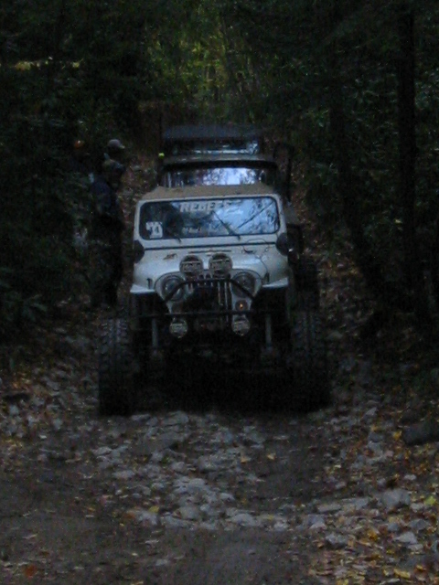 1996 Ford Bronco - A white Jeep CJ-7 navigates a rocky, wooded path, with another vehicle visible behind it.