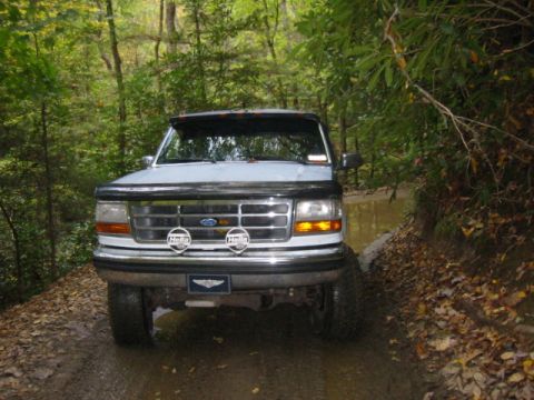 1996 Ford Bronco - This 1996 Ford Bronco navigates a wet, wooded path, its front grille and headlights clearly visible.