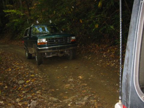 1996 Ford Bronco - A green Ford Bronco drives on a dirt road surrounded by trees, seen from a 3/4 front angle.