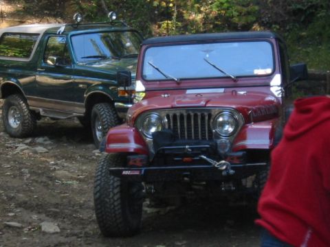 1996 Ford Bronco - Two off-road vehicles, a red Jeep and a green Ford Bronco, are parked on a dirt path surrounded by trees.
