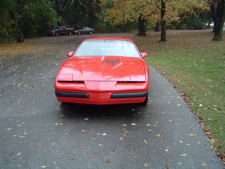 Front view of a red Pontiac Firebird parked on a leaf-strewn driveway with pop-up headlights closed