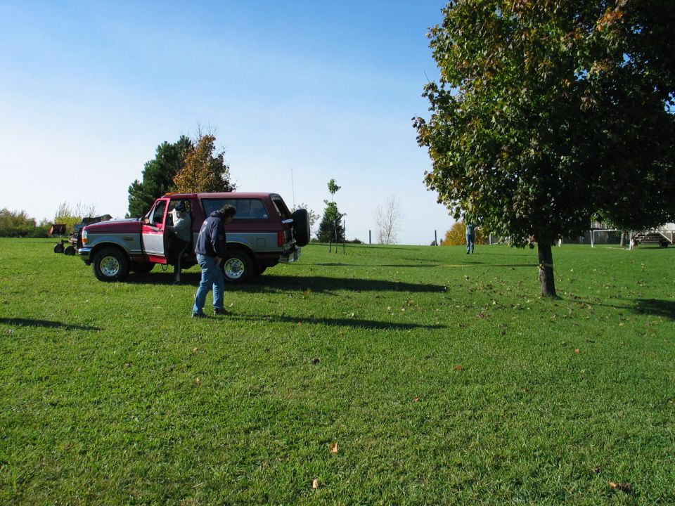 Red and gray SUV parked on grass, two people near the open driver's door. Another person is walking in the background