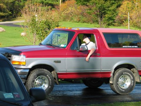 1995 Ford Bronco - A red and silver 1995 Ford Bronco is seen from the side, with a driver visible through the window.