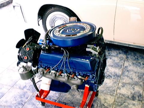 1979 Ford Bronco - A blue engine is displayed on a red stand, partially obscuring a white car's fender and wheel.
