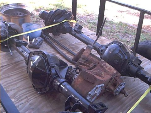 1979 Ford Bronco - Several vehicle axles and a rusty transmission are laid out on a wooden surface, possibly for sale or repair.