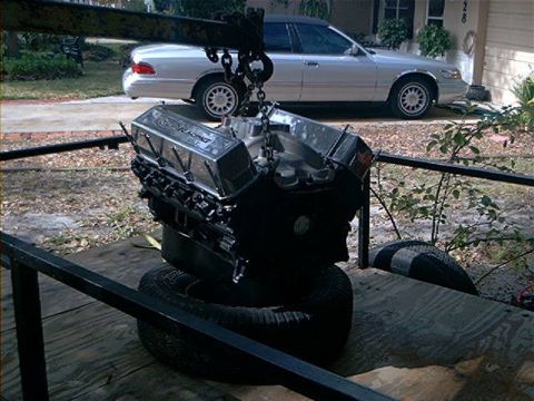 1979 Ford Bronco - A Ford Racing engine is hoisted by a crane, with a sedan in the background.