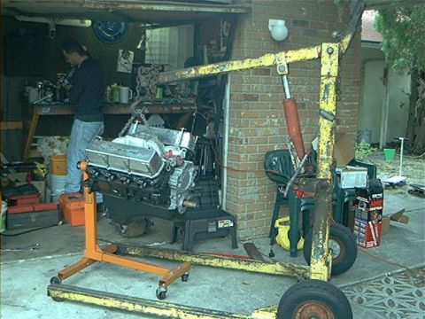 1979 Ford Bronco - A person works on an engine that is suspended by a yellow engine hoist in a cluttered garage.