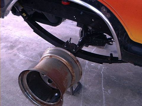 1979 Ford Bronco - A detached rusty wheel rests on the ground near the undercarriage of a vehicle, showing leaf spring suspension.