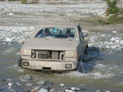 1985 Ford Bronco - This pickup truck navigates a rocky riverbed, with two occupants visible inside the cab.