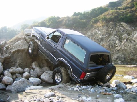 1985 Ford Bronco - A dark blue SUV with chrome trim is shown navigating a challenging rocky terrain next to a body of water.