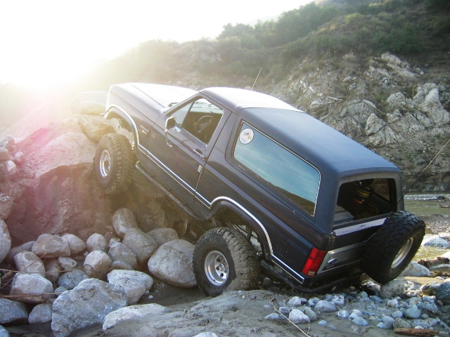 Dark blue Ford Bronco SUV climbing boulders off-road on large tires in a rocky riverbed.