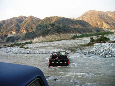 1985 Ford Bronco - A black Ford pickup truck navigates a shallow river, with a man sitting in the open bed as mountains loom in the distance.