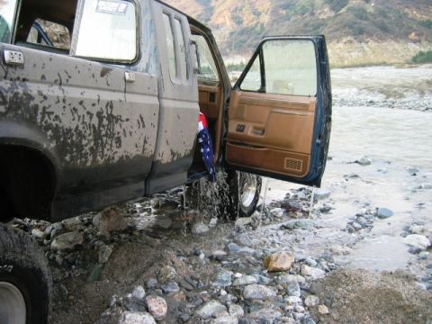 1985 Ford Bronco - A muddy 1987 Ford F-150 pickup truck is stuck in a rocky stream with its door ajar and an American flag visible inside.